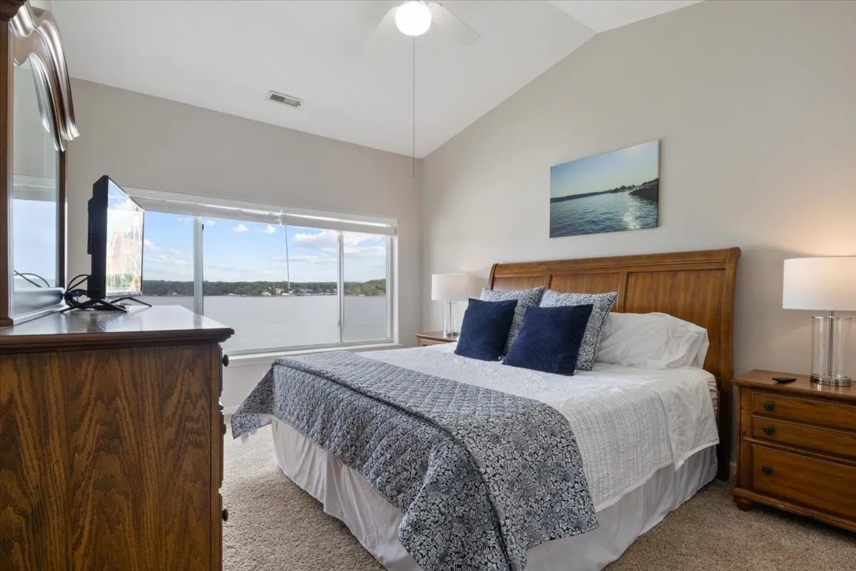 This image showcases a serene bedroom featuring a bed with decorative pillows, a large window offering a beautiful lake view, and a television placed on a dresser. The space feels calm and peaceful, ideal for relaxation.