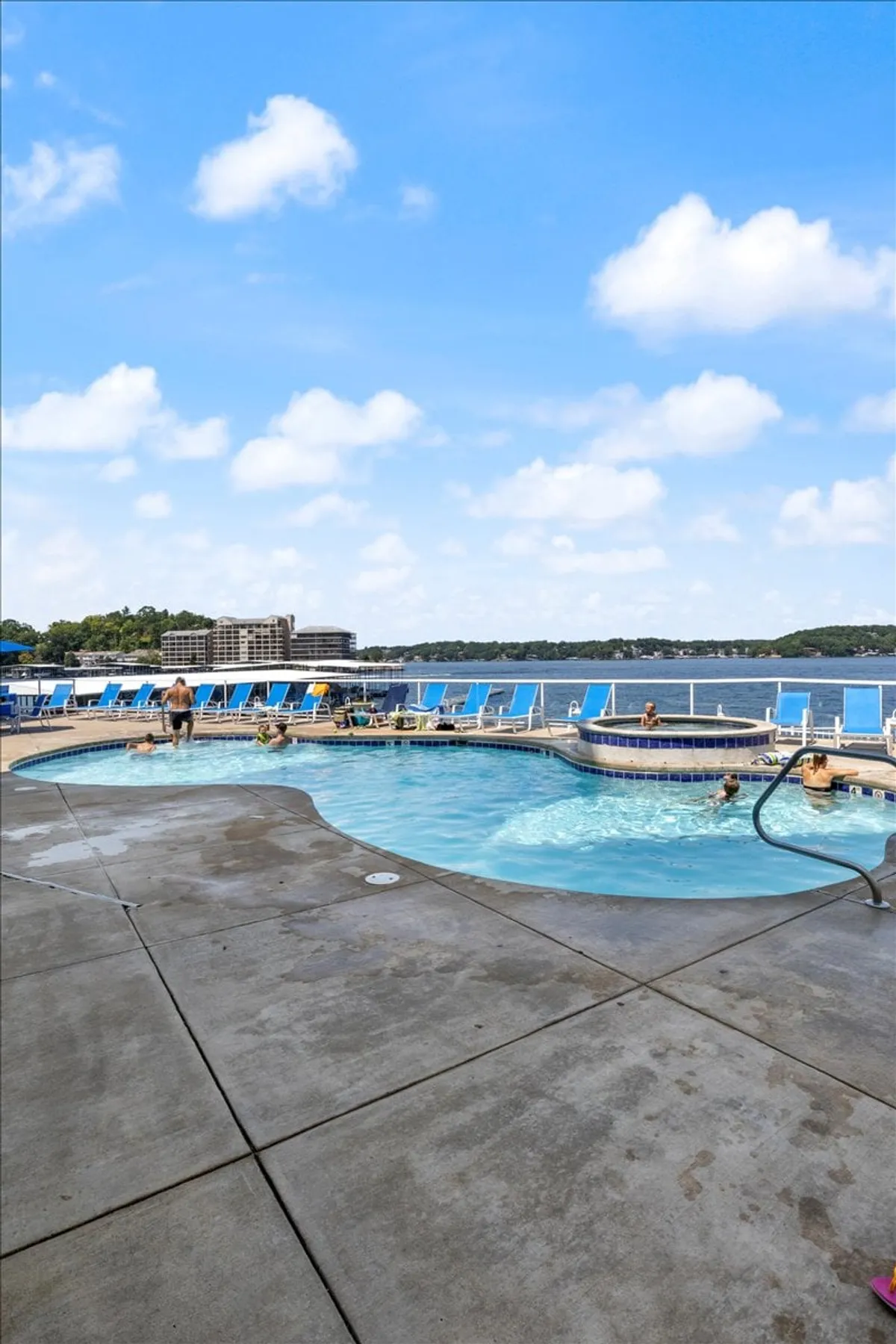 This image highlights an outdoor pool area with both a swimming pool and hot tub, surrounded by lounge chairs and overlooking the lake. The atmosphere is bright and friendly, perfect for social gatherings or relaxing by the water.