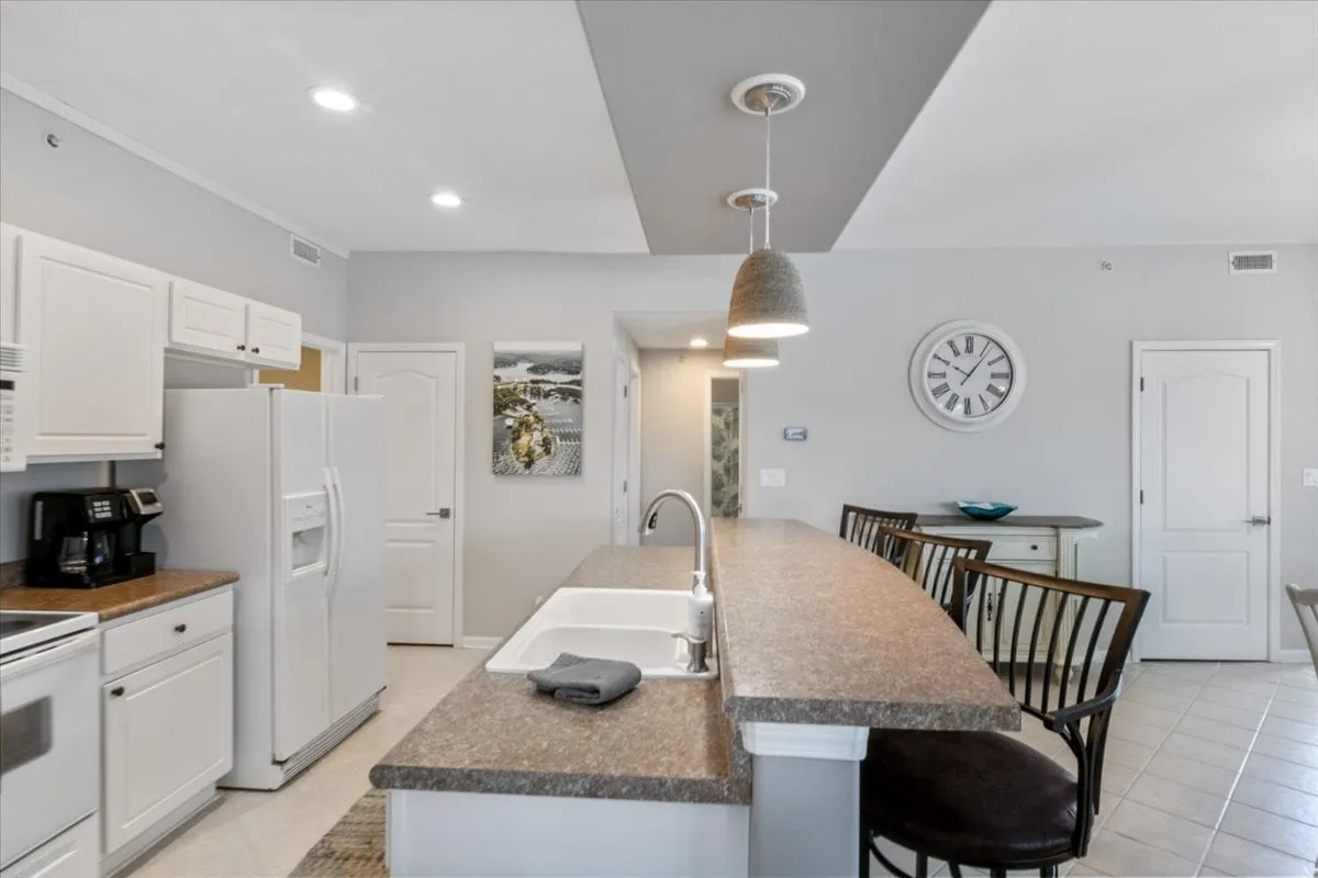 A warm, open kitchen scene featuring a large kitchen island with bar stools, white cabinetry, and a wall clock. The island invites casual dining or gathering for a quick breakfast, while the layout gives a full view of the kitchen’s functional design.