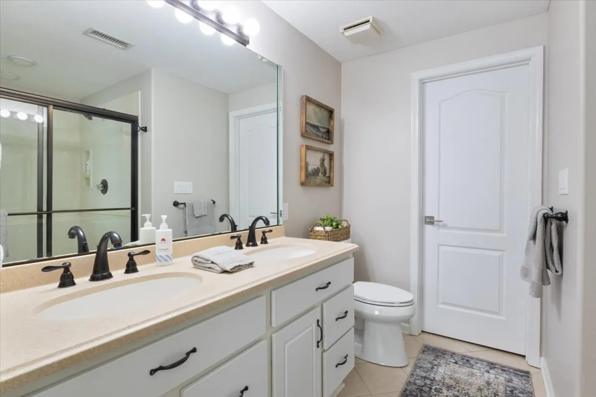 A spacious bathroom with double sinks, a large mirror, framed artwork, and elegant light fixtures. The atmosphere is inviting and functional, perfect for morning and evening routines.