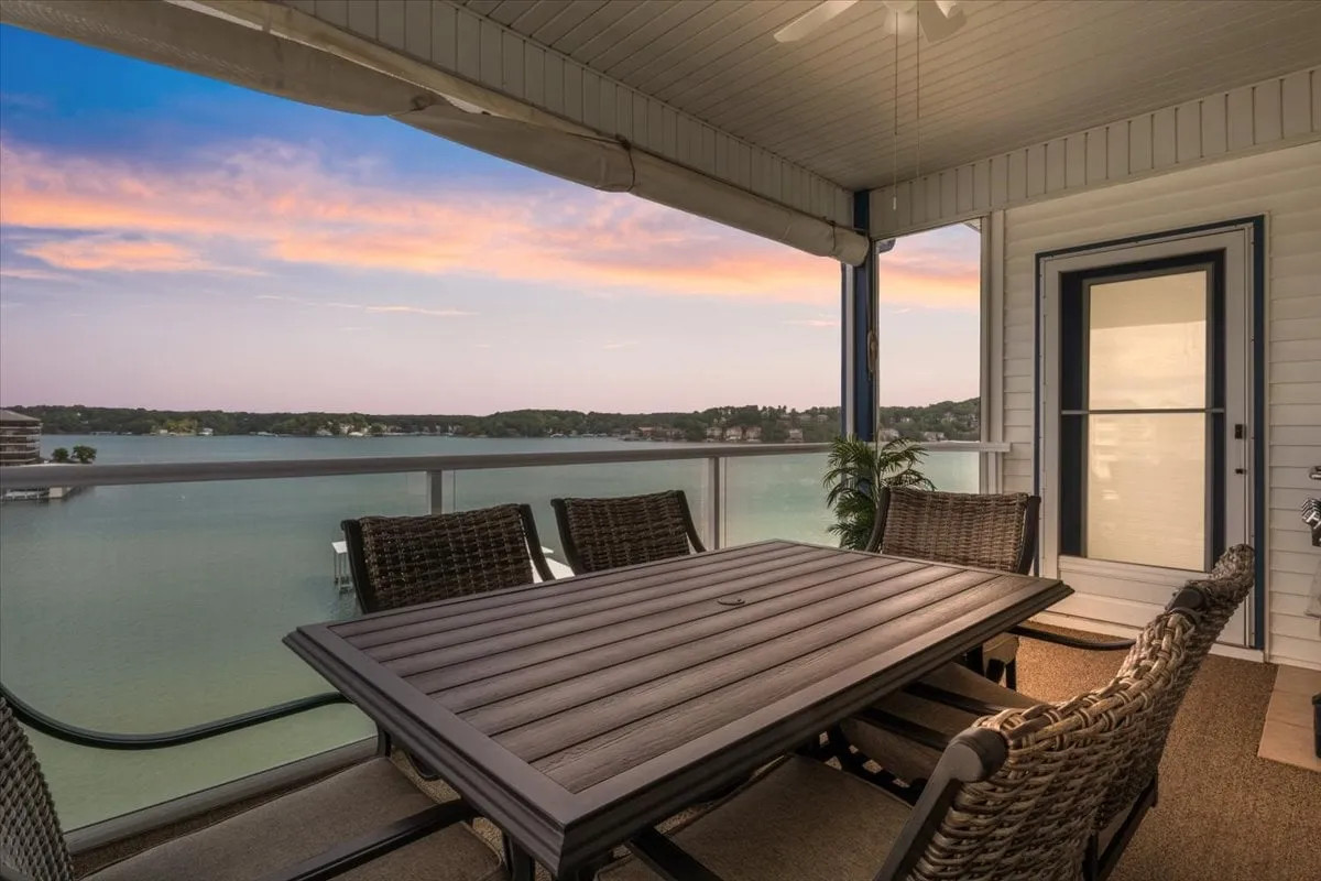 The image showcases a serene outdoor dining area set on a screened porch, with comfortable chairs arranged around a dining table. Beyond the porch, there is a sweeping, unobstructed view of Lake Ozark’s sparkling waters under a sky painted with sunset hues, creating an inviting space for relaxation and social gatherings.