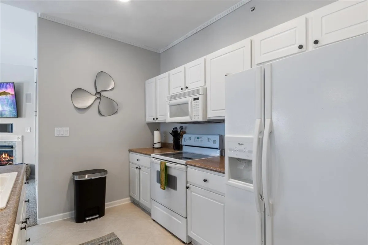This image features a modern kitchen with bright white cabinetry, sleek granite countertops, and stainless steel appliances. The space is well-lit and welcoming, with decorative touches like a wall fan adding personality. Ample workspace and visible cookware suggest it's ready for group meal preparation.