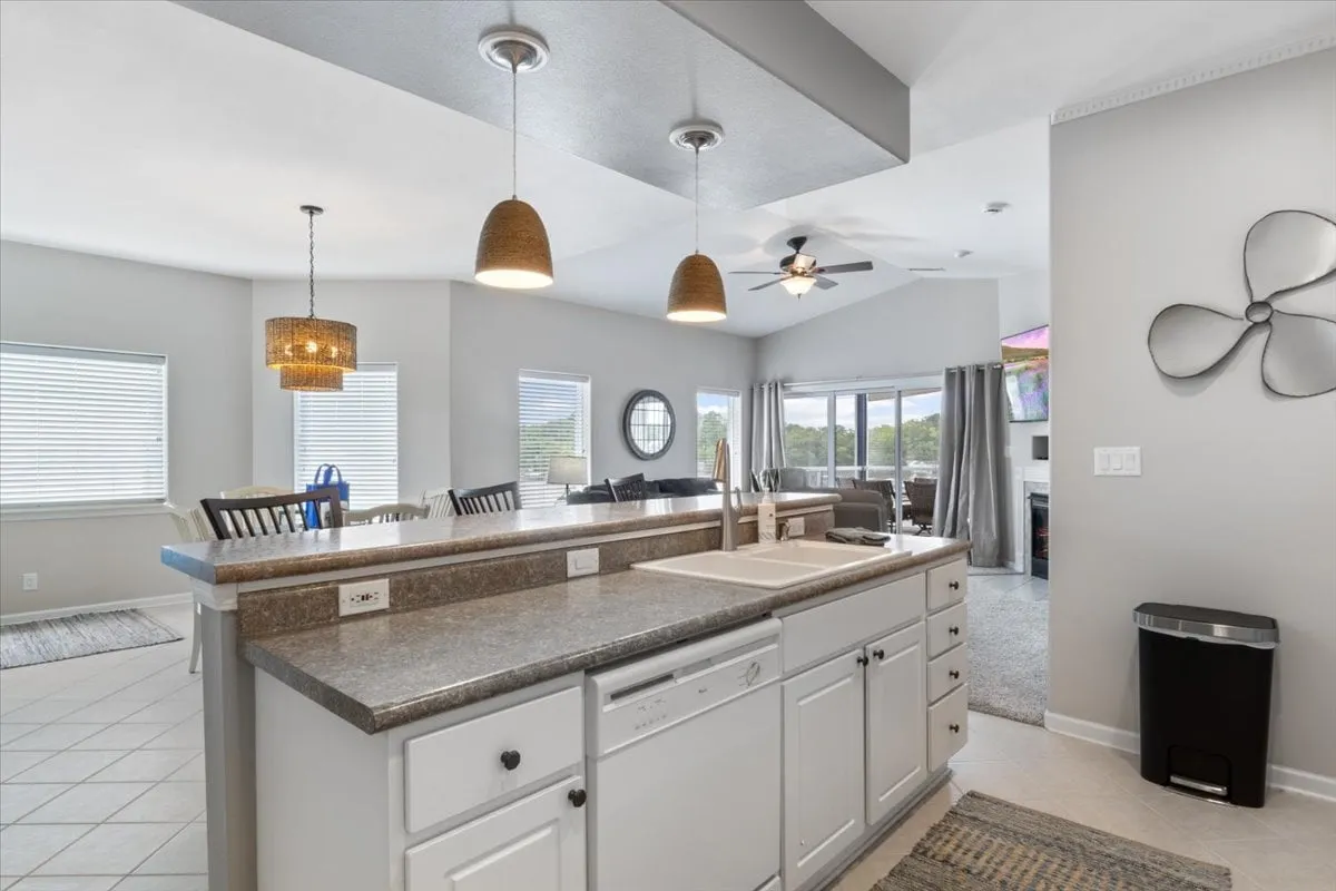 This image captures a bright, open-concept space featuring a kitchen island, pendant lights, a spacious dining area, large windows, and an adjacent living area. The cozy, inviting atmosphere is accentuated by comfortable seating and expansive views through the windows, suggesting a seamless flow between living, dining, and entertaining spaces.
