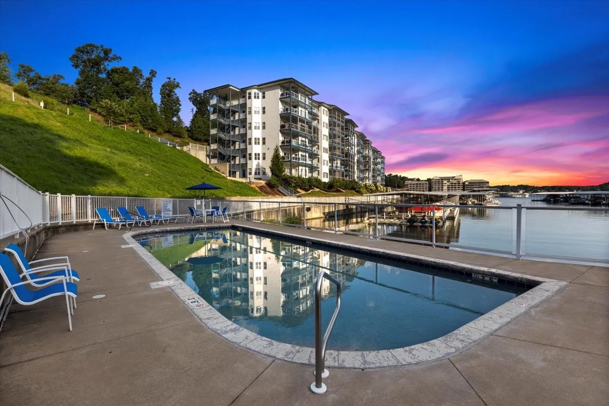 A stylish swimming pool overlooks the lake, with the waterfront view framed by a modern apartment building and gentle hills. The scene is serene and inviting, hinting at the resort-style relaxation available after a day on the water, whether swimming, sunbathing, or socializing by the pool.