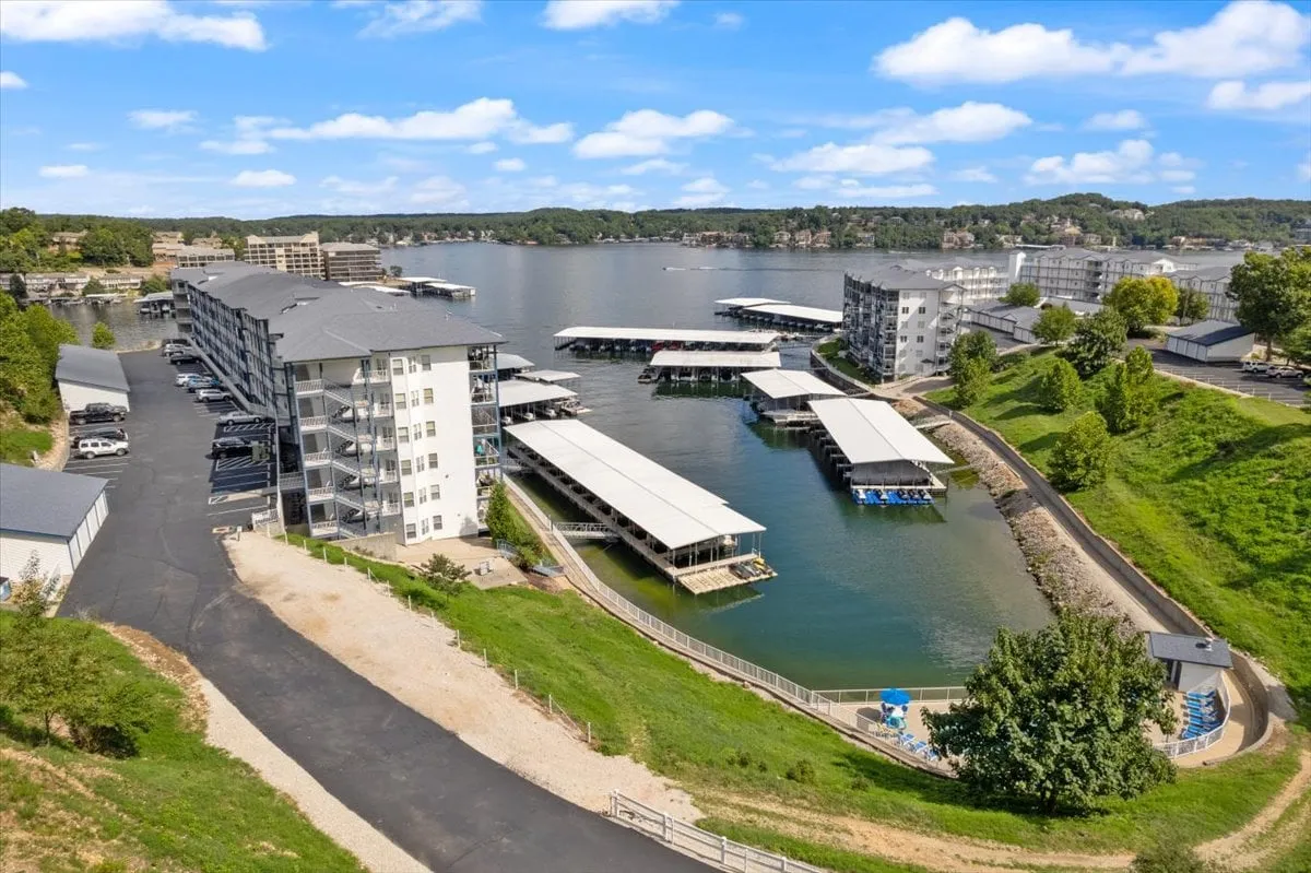The image shows a multi-story building adjacent to a marina, with boats docked along the shoreline and greenery surrounding the area. The view highlights both the proximity to watercraft activity and the relaxed lakeside atmosphere. It also indicates the presence of local marinas, connecting guests to rental and boating options.