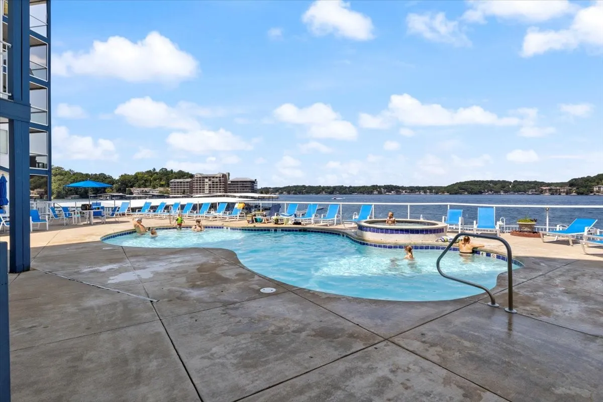 This image displays a vibrant outdoor pool area featuring a sparkling swimming pool, a rejuvenating hot tub, lounge chairs, and a scenic lake view. The setting is lively and cheerful, capturing people relaxing, swimming, and socializing under the sun.